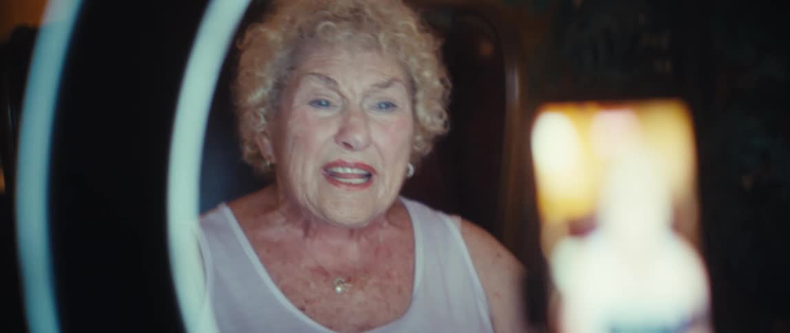 An elderly woman talks into a smart phone while holding a chart in her hand and a ring light around her