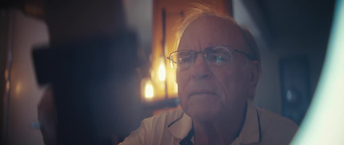 An elderly man wearing glasses with a look of serious concentration as he is brightly illuminated by a ring light while operating a smartphone mounted on the ring light setup