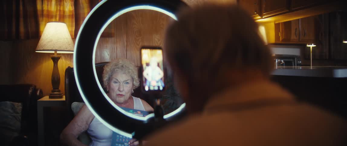 An elderly woman sits behind a ring light in the living room live streaming while a man behind the camera monitors the process