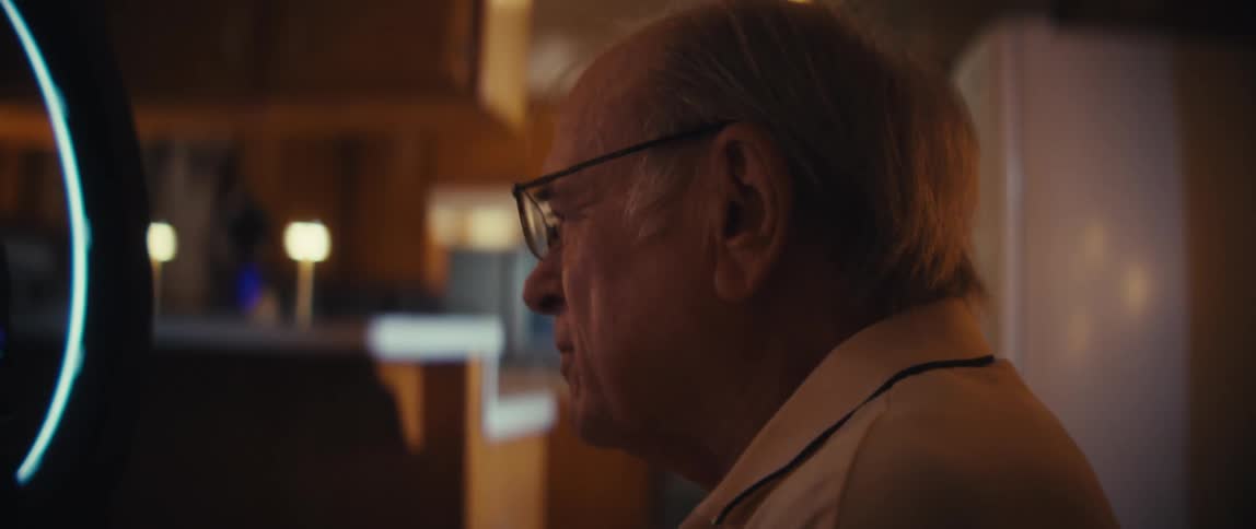 An elderly man wearing glasses reveals him looking intently at a smartphone screen which is mounted on a stand and illuminated by a ring light showing a fullscreen video of a woman sitting in front of the ring light