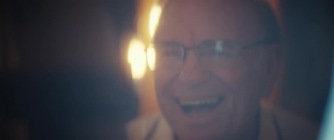 An elderly man wearing glasses records with his smartphone indoors illuminated by a ring light creating excitement
