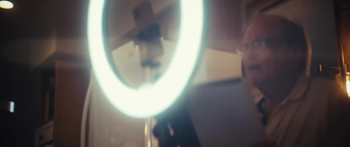 The clip features two senior individuals in an interior setting likely filming or taking a photograph using a phone mounted in a large ring light the woman with curly blonde hair initially smiles and then the focus shifts to the man wearing glasses