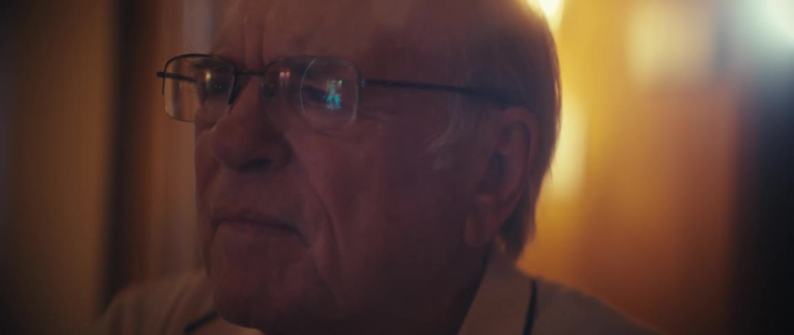 An elderly man wearing glasses sits indoors scrolling on a mobile device