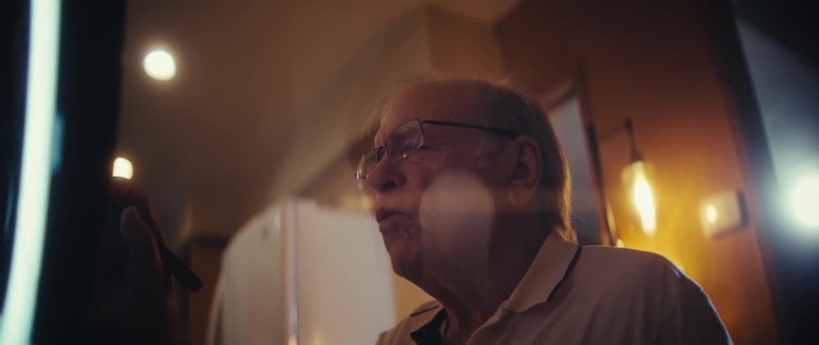 An elderly man leans in to speak to his smartphone his face illuminated by the bright glow of a ring light as he records himself in a warm room