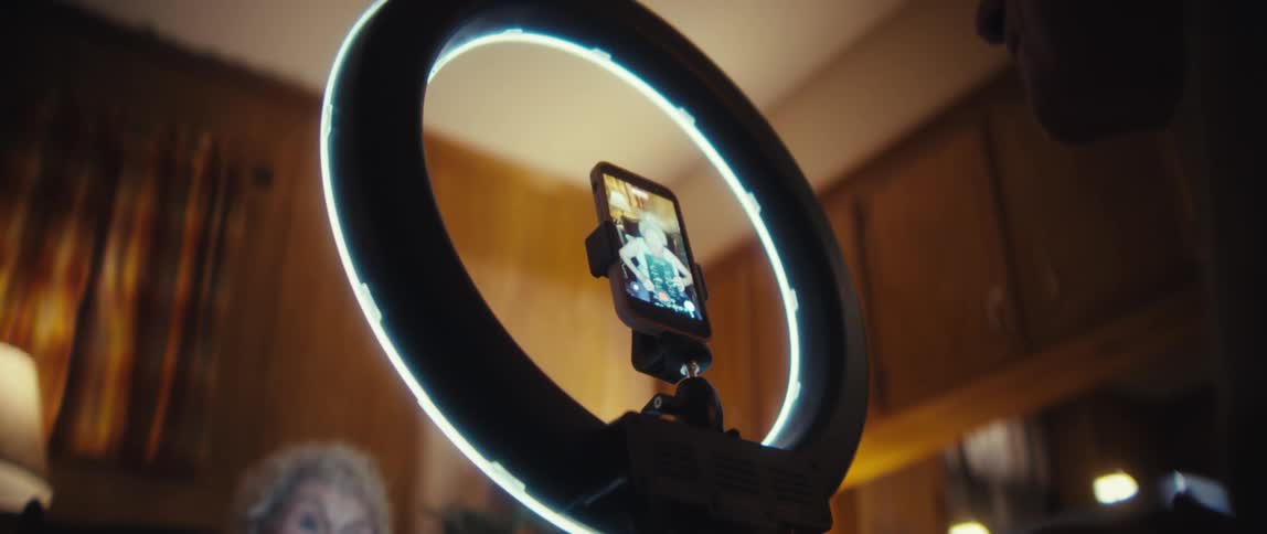 Woman holding a printed sheet of paper while recording a video in front of a ring light which her husband is watching