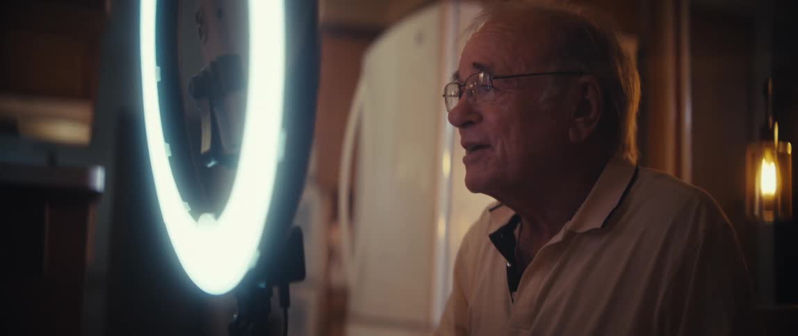 This closeup video shows an older man wearing glasses and a lightcolored polo shirt seated indoors and looking offcamerahe is being lit from the front by a large circular ring light with a smartphone mounted in the center suggesting he is record