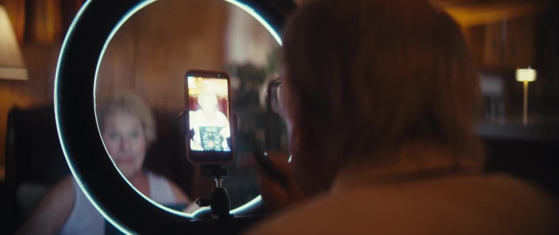 A elderly man faces a phone mounted on a stand inside a bright circular ring light and uses a finger to interact with the phones screen while pointing the phone to a elderly woman on the back