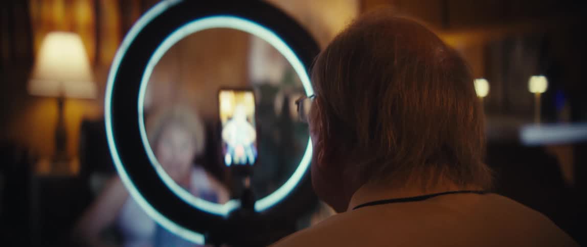 Grayhaired person films elderly woman lit by ring light in cozy room with wooden walls