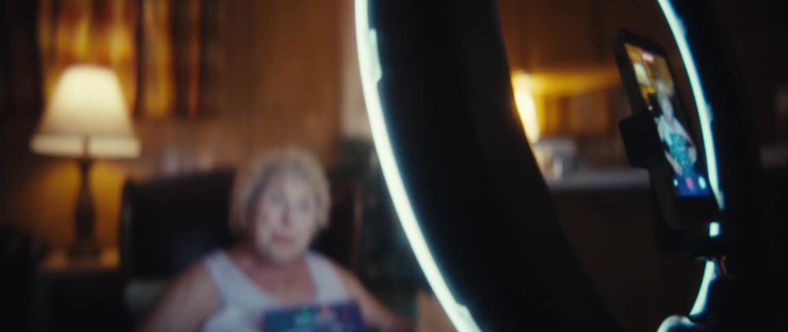 The video clip shows a closeup of a smartphone screen attached to a ring light displaying a video recording of an elderly womanthe camera then racks focus to the woman who is sitting in a dark cozy room holding a graphic chart titled trading can