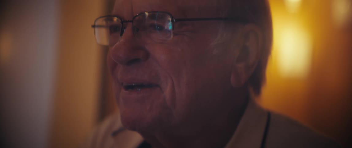 An elderly man wearing glasses sits in his living room watching a computer screen with excitement