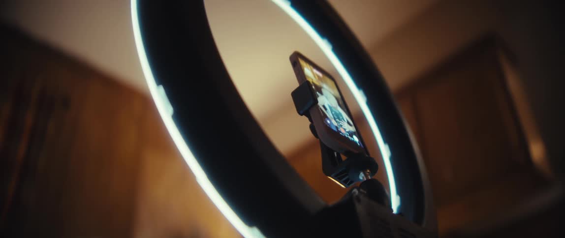 The video shows a closeup profile view of an older man wearing glasses smiling slightly while looking at a smartphone screen mounted in a ring light the camera then focuses on the ring light and the phone before panning down to reveal an older woman wi