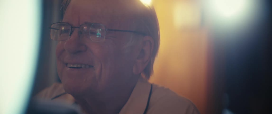Man with glasses and light shirt writes in notebook under soft light in indoor setting