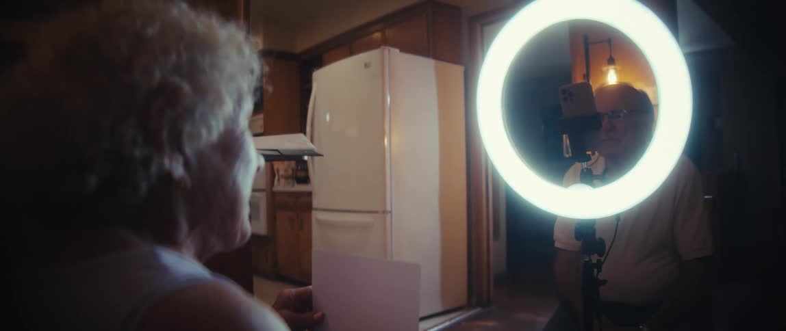 An elderly man helping his female partner record a mobile video indoors in the dining area