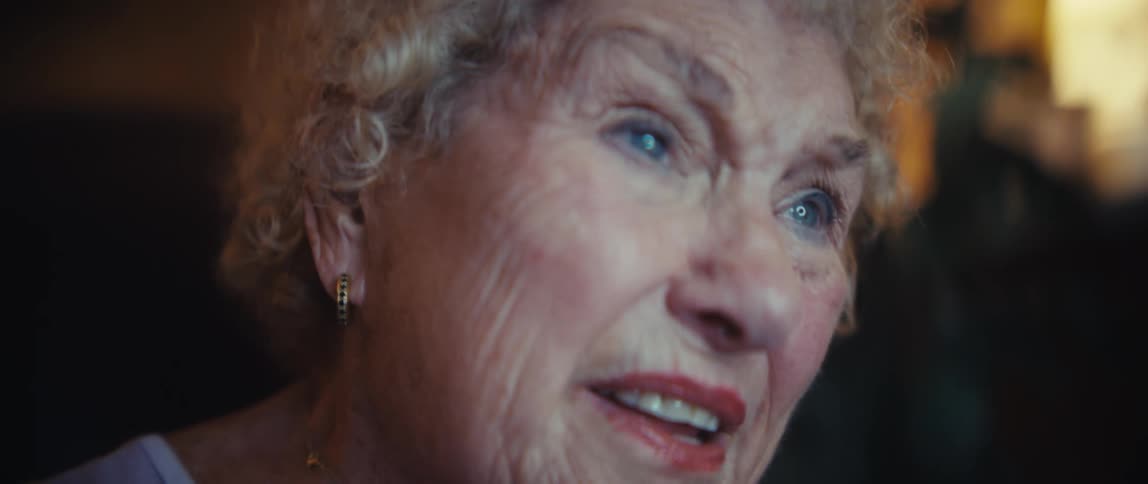 A closeup of an elderly womans hand holding a darkcolored book as she looks up and speaks with an engaged expression in an indoor setting