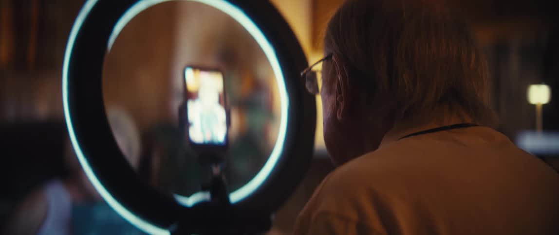 Man adjusting smartphone plugged into ring light while recording an elderly woman indoors