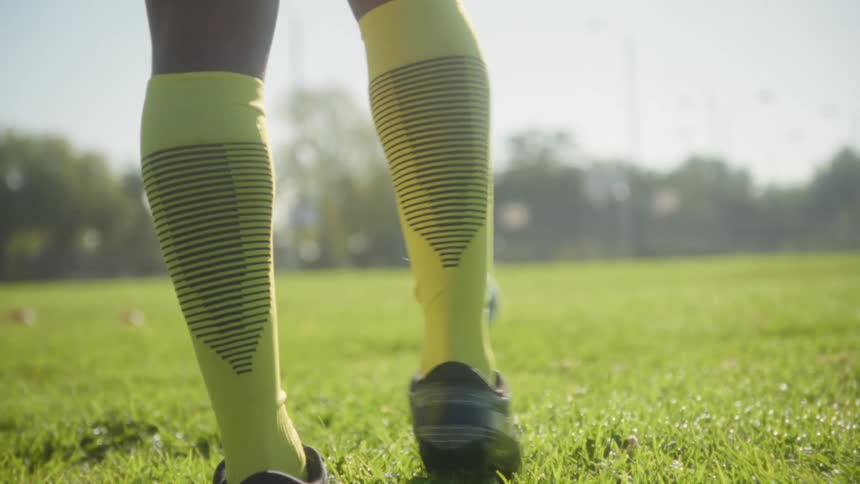 A lowangle closeup shot captures the legs of a soccer player wearing yellow high socks and cleats as they step across a sunny green grass field during practice