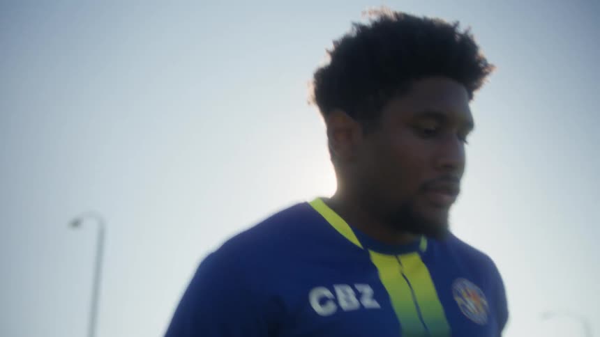 A closeup shot angled low shows a soccer player wearing a blue and yellow jersey running forward in an open field against the bright clear sky