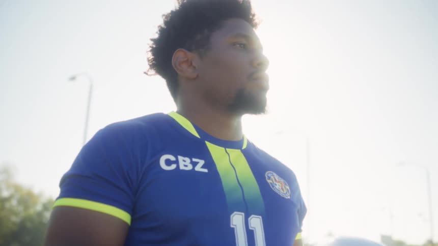 The video shows a young man wearing a blue soccer jersey with yellow and green trim marked with the number 11 and cbz he holds a black and white soccer ball in his hands the camera focuses on him as he looks thoughtfully or determinedly offscreen in