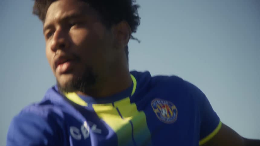 A male soccer player wearing a blue and yellow jersey with the number eleven shows him in the midst of intense physical action his face exhibiting a focused and determined expression against a bright clear blue sky