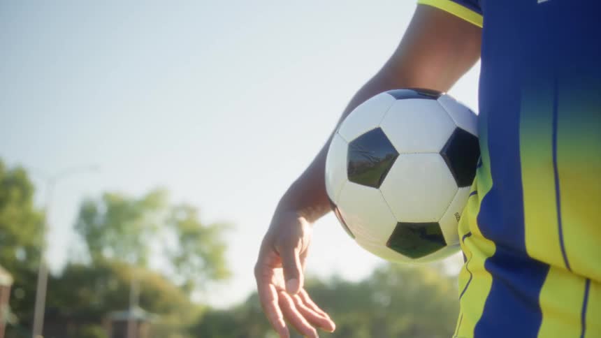 Player in blue uniform with yellow accents holds classic ball against body under clear sky
