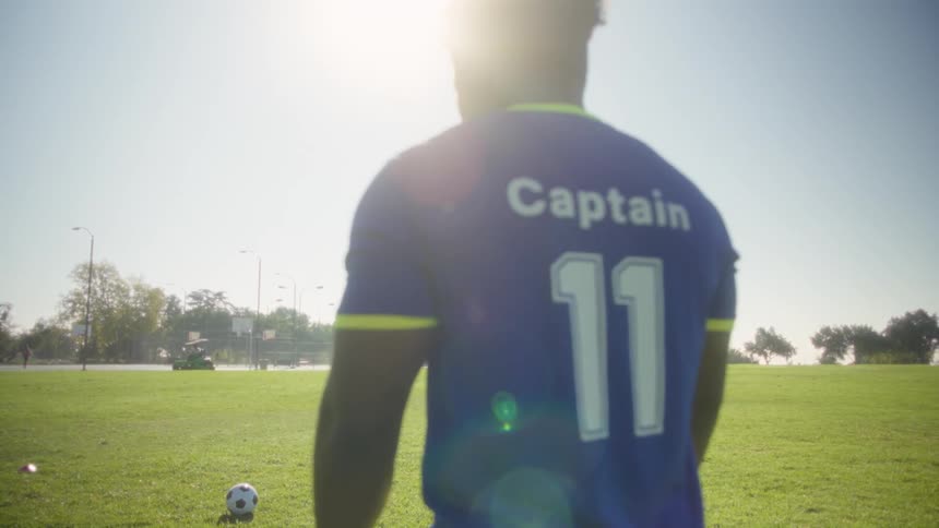 A medium shot shows a male soccer player from behind wearing a blue jersey standing near a soccer ball on a sunny grass field before a strong bright backlight