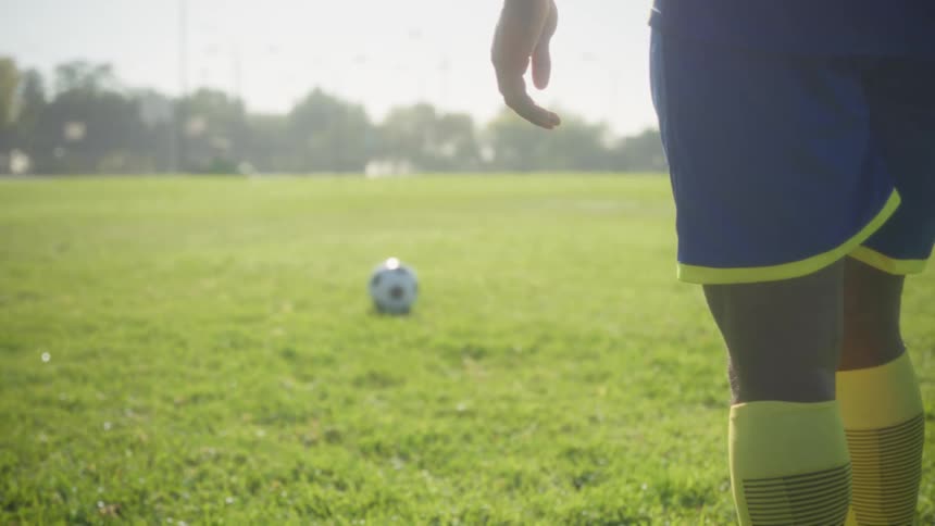 A soccer player wearing blue shorts and yellow socks with their right hand dangling at their side and a soccer ball resting on the bright green field