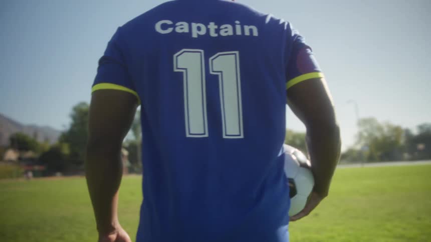 Soccer player walking across the field wearing a captains jersey and holding a ball