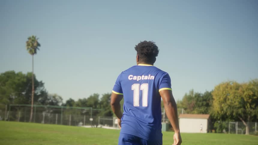 A man wearing a blue soccer jersey with the word captain and the number 11 on the back walks across a sunny green fieldthe camera follows him from behind as he heads toward a distant white building and trees under a clear blue sky