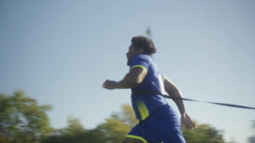 A male soccer player in a blue and yellow uniform runs across a sunny green field pulling a resistance parachute with mountains and trees in the background