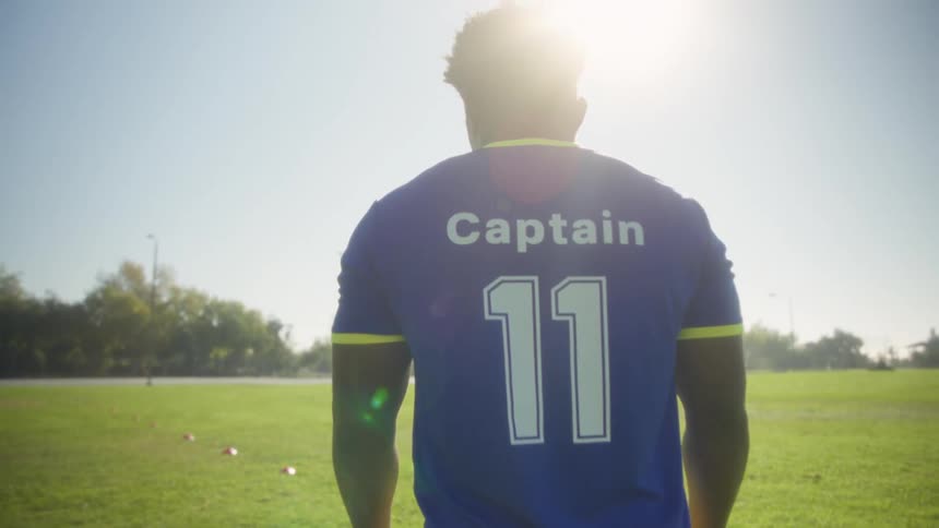 A young black male stands on a football field on a sunny day wearing a blue jersey with 11 and captain written on the back