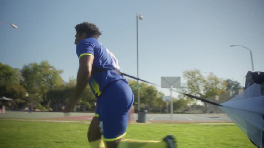 A soccer player wearing a blue and yellow uniform runs rapidly across a grassy field under a bright sky pulling a deployed resistance parachute behind him with a mountain range visible in the distant background