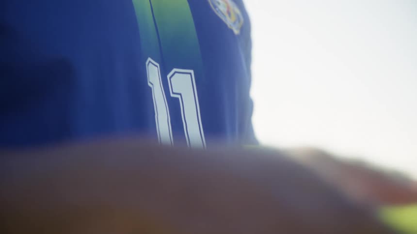 The closeup shot transitions from the profile of a soccer players face heavily backlit by bright sunlight to a focus on his jersey and finally shifts to his hands as he holds a traditional black and white soccer ball on a green outdoor field