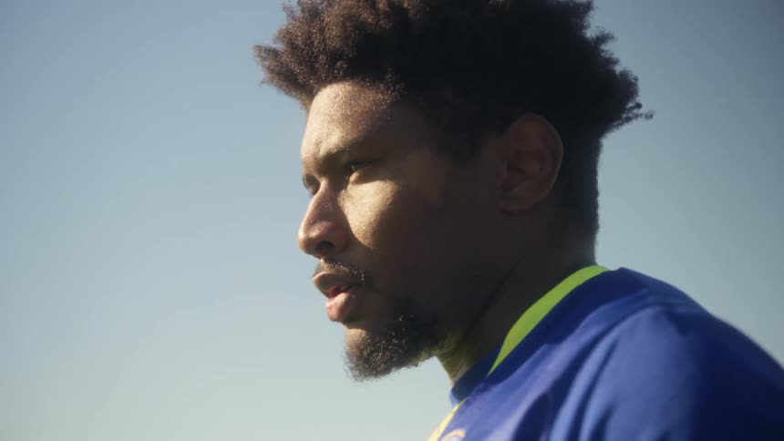 A tight closeup shows the profile of a soccer player wearing a blue and yellow jersey with intense focus on his face and determined mouth as he looks offcamera against a bright blue sky