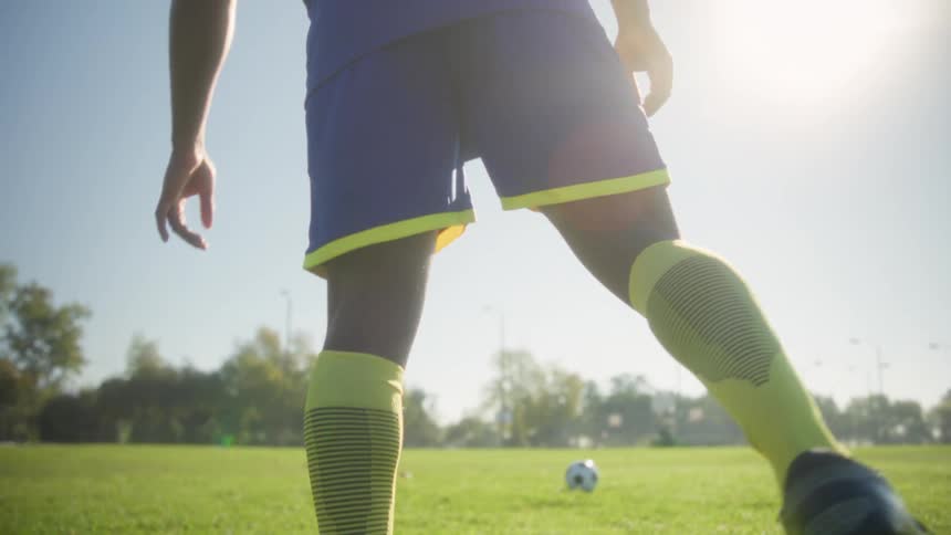 This lowangle shot begins by focusing on a soccer players legs in bright yellow socks and cleats stepping toward a ball on a sunny grass fieldthe camera then pans up to show the players back wearing a blue jersey marked captain 11 dramatically