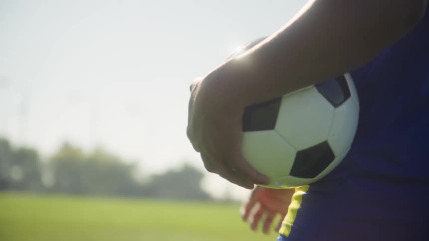A male carries a soccer ball in his hand while walking on a soccer field