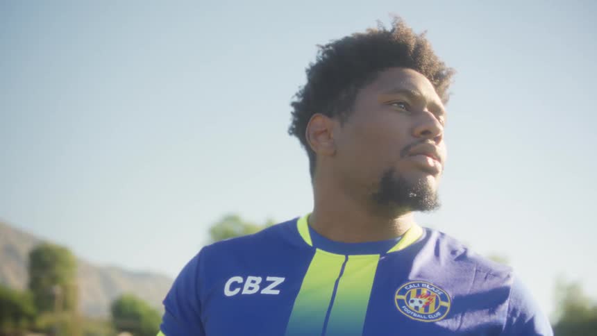 A male soccer player with dark curly hair and a beard wearing a blue and yellow jersey and holding a soccer ball looks intently into the distance in an outdoor sunlit field with mountains in the background