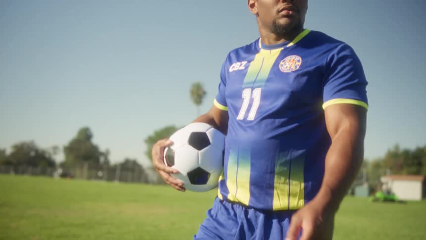 Player in blue uniform with green and yellow stripes holds ball on grassy field under clear sky