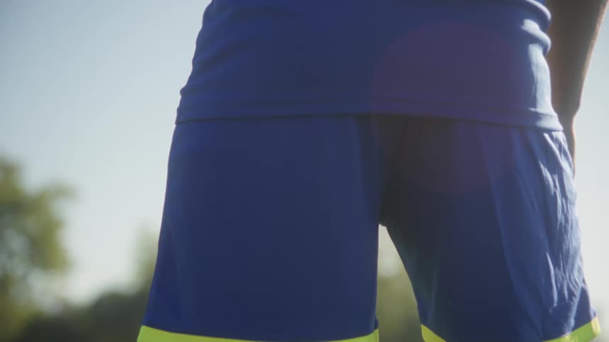 The video begins with an overtheshoulder shot of a male soccer players back wearing a blue jersey with captain and the number 11 facing bright sunlight the camera slowly pans down to a lowangle closeup of his legs in blue shorts and bright yello