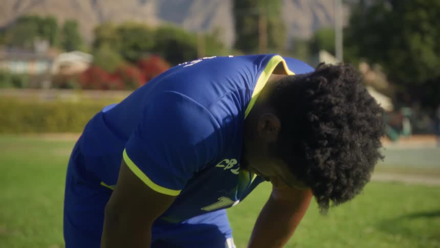 An exhausted soccer player runs his hand through his hair before bending over to catch his breath on a sunny field