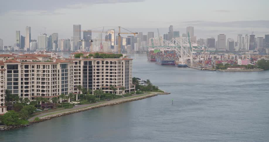An elevated view shows a luxury residential building on the waterfront overlooking a dense city skyline and a busy commercial port with cranes on a hazy day