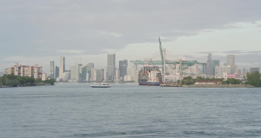 This is a wide shot of a busy urban waterfront on a slightly overcast day likely showing the port of a major city like miami the foreground is dominated by choppy blue water with a small white boat moving across the channel