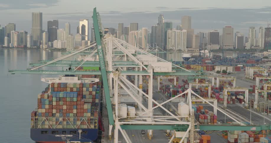 An aerial view shows a busy shipping yard with stacked containers and cranes framed by a distant city skyline across the water