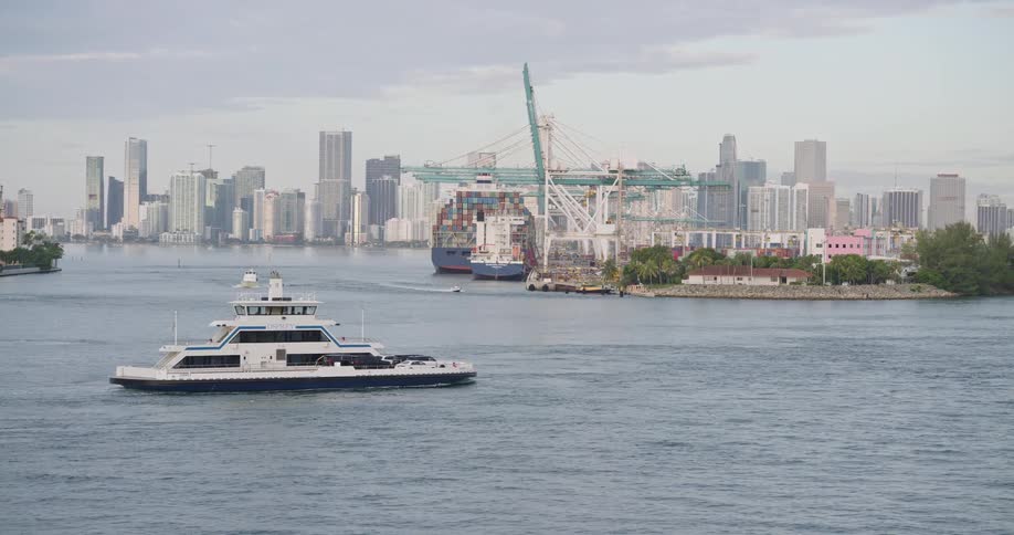 A boat rides through ocean waters near the coastline at midday passing a seaport where containers are docked with the city skyline visible in the background