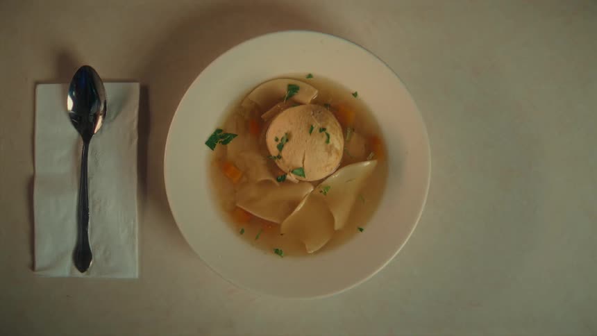 A hand sets a white bowl of clear broth soup with large matzo ball and wide noodles down onto a table next to a spoon on a white napkin