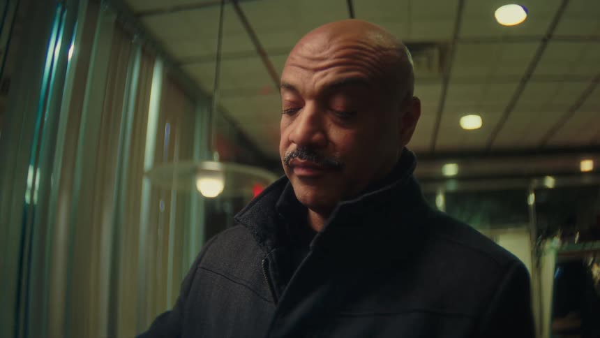 A middleaged bald man with a mustache wearing a dark jacket is captured in a medium closeup shot looking downward and filmed in an interior location with patterned ceilings and vertical curtains visible behind him