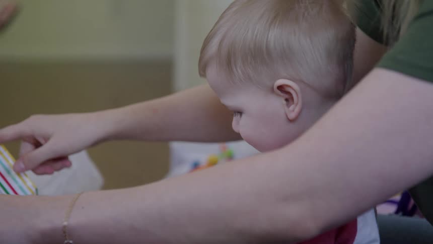 A toddler with blonde hair sits closely next to an adult playing with a small board book while the adults hands help him to get a new toy in an indoor setting