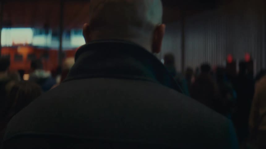 A bald man in a dark coat walks through a crowded indoor terminal