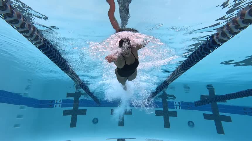 An underwater view shows a woman in a black swimsuit and cap swimming the freestyle stroke quickly down a clear blue pool lane