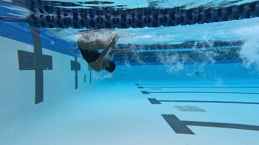 An underwater view shows a woman in a black swimsuit and cap performing a flip turn and pushing off the wall in a clear blue swimming pool