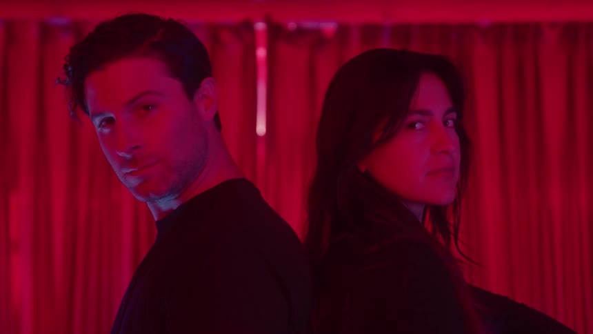 A man and a woman in dark clothing stand back to back turning their faces toward the camera in a room bathed in dramatic red lighting